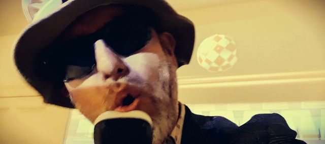 die nmi singing into a microphone, wearing a hat and sunglasses. The picture has a lot of dynamic blur.